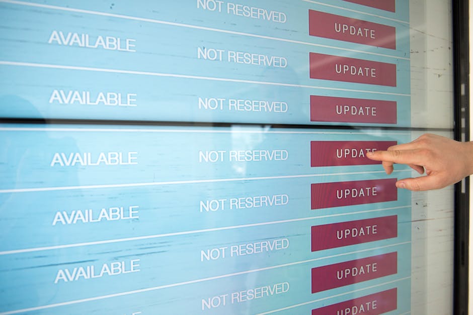 A person’s hand pointing at a digital screen displaying multiple rows with the words 'AVAILABLE' and 'NOT RESERVED' in white text, alongside red 'UPDATE' buttons. The background includes a reflection 